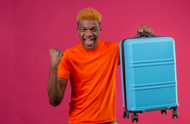 Довольный молодой красивый мальчик в оранжевой футболке с дорожным чемоданом