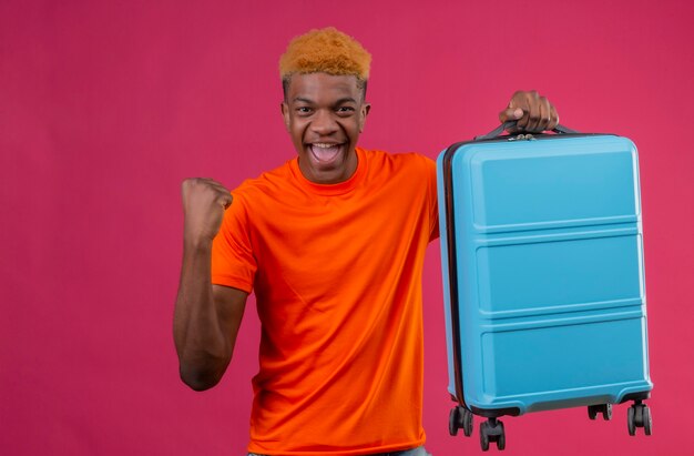 Pleased young handsome boy wearing orange t-shirt holding travel suitcase