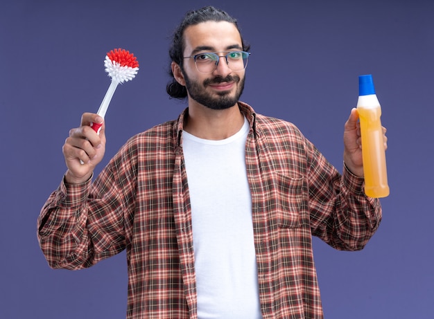Felice giovane bel ragazzo delle pulizie indossando t-shirt tenendo la spazzola con detergente isolato sulla parete blu