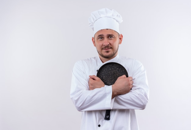 Pleased young handsome cook in chef uniform holding frying pan isolated on white space