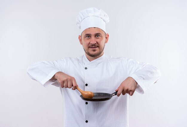 Pleased young handsome cook in chef uniform holding frying pan and spoon isolated on white space