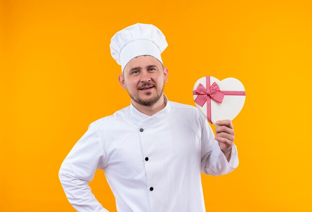 Pleased young handsome cook in chef uniform holding heart shaped gift box and raising finger on isolated orange space