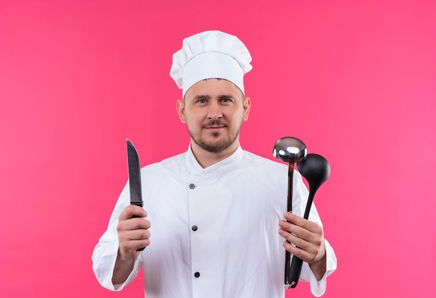 Pleased young handsome cook in chef uniform holding knife and ladles isolated on pink space