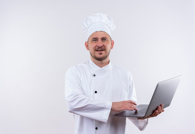 Pleased young handsome cook in chef uniform holding laptop on isolated white space 