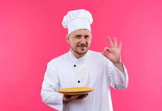 Довольный молодой красивый повар в униформе шеф-повара держит тарелку и делает знак ОК, изолированные на розовом пространстве