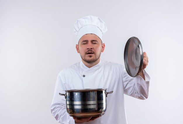 Pleased young handsome cook in chef uniform holding pot and pot lid with closed eyes isolated on white space