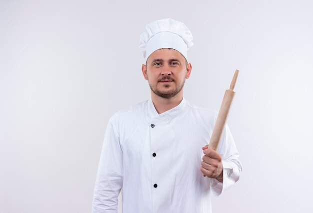 Pleased young handsome cook in chef uniform holding rolling pin isolated on white space
