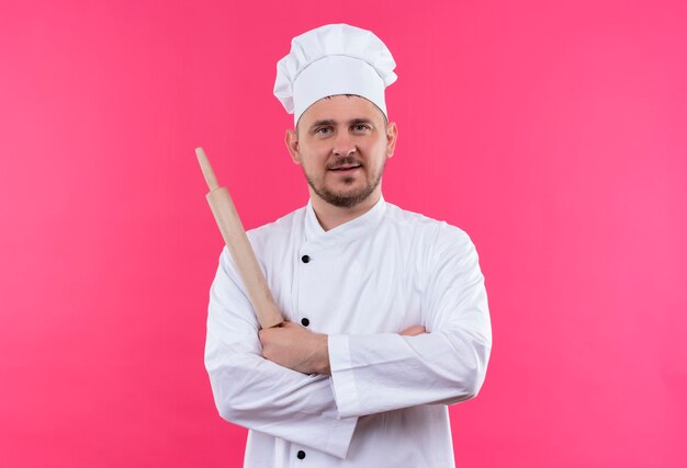 Pleased young handsome cook in chef uniform holding rolling pin and standing with closed posture isolated on pink space