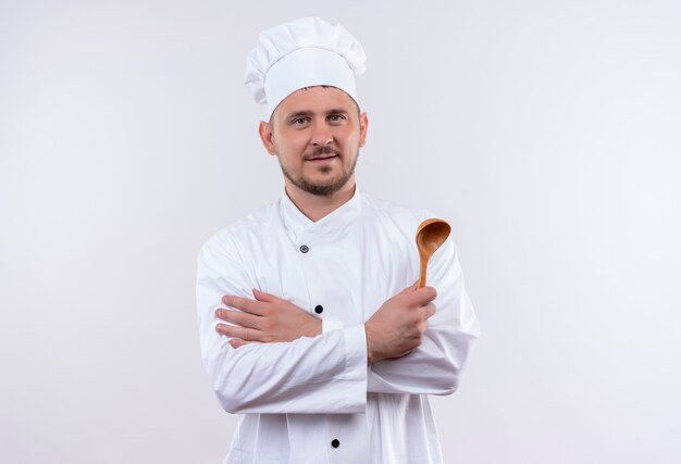 Pleased young handsome cook in chef uniform holding spoon standing with closed posture isolated on white space