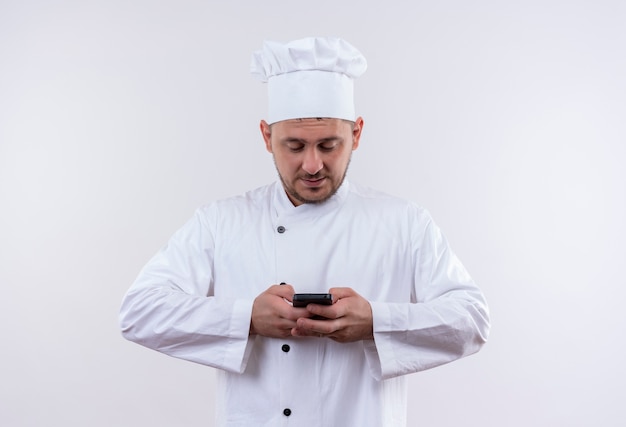 Pleased young handsome cook in chef uniform holding and using mobile phone isolated on white space