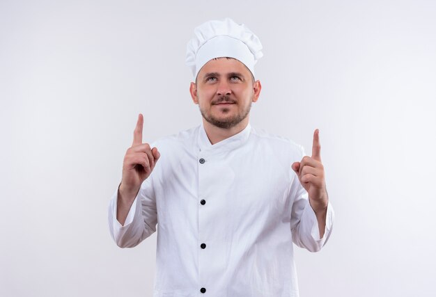 Pleased young handsome cook in chef uniform pointing and looking up isolated on white space