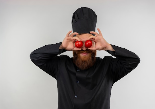 Pleased young handsome cook in chef uniform putting tomatoes on eyes isolated on white space