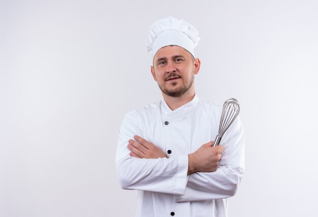 Pleased young handsome cook in chef uniform standing with closed posture and holding whisk isolated on white space