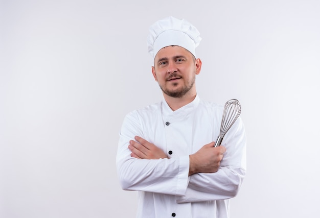 Pleased young handsome cook in chef uniform standing with closed posture and holding whisk isolated on white space