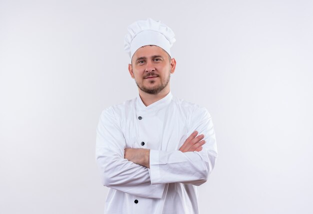Pleased young handsome cook in chef uniform standing with closed posture isolated on white space
