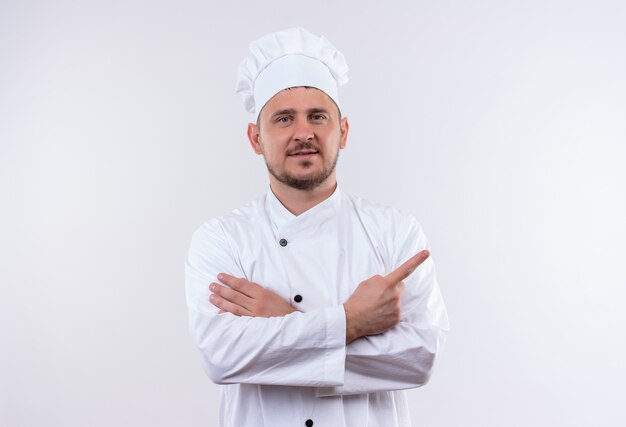 白いスペースで隔離の右側を指している閉じた姿勢で立っているシェフの制服を着た若いハンサムな料理人を喜ばせる