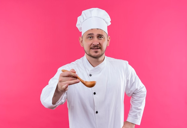 Pleased young handsome cook in chef uniform stretching out spoon isolated on pink space