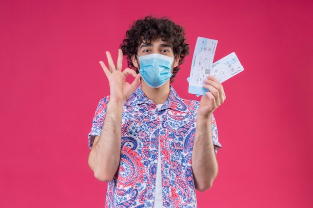 pleased young handsome curly traveler man wearing medical mask holding airplane tickets doing ok sign on isolated pink space with copy space