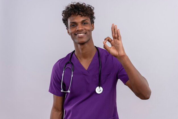 A pleased young handsome dark-skinned doctor with curly hair wearing violet uniform with stethoscope showing ok sign with fingers 