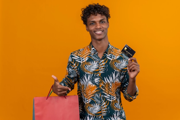 Pleased young handsome dark-skinned man with curly hair in leaves printed shirt smilingholding shopping bags showing credit card while standing on an orange background