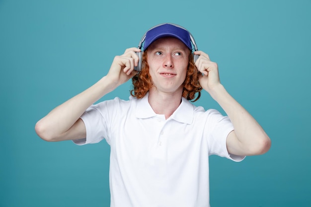 Pleased young handsome guy in cap wearing headphones isolated on blue background