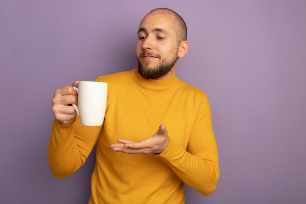 Soddisfatto giovane ragazzo bello che tiene e indica con la mano alla tazza di tè isolata sulla parete viola con lo spazio della copia