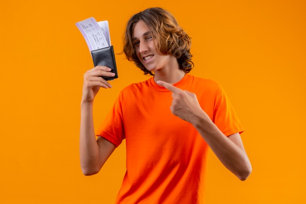 自信を持って笑顔でカメラを見て彼らに指で指している航空券を保持しているオレンジ色のtシャツで満足している若いハンサムな男