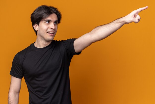 Pleased young handsome guy wearing black t-shirt points at side isolated on orange wall