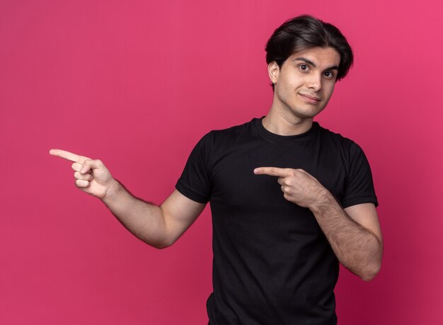 Pleased young handsome guy wearing black t-shirt points at side isolated on pink wall with copy space