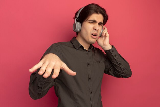 Pleased young handsome guy wearing black t-shirt with headphones showing dj gesture isolated on pink wall