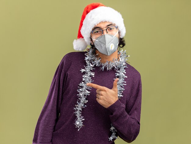 Pleased young handsome guy wearing christmas hat and medical mask with garland on neck points at side isolated on olive green background with copy space