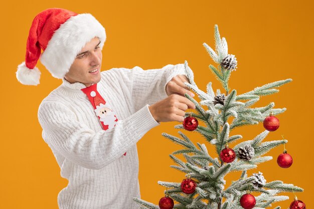 pleased young handsome guy wearing christmas hat and santa claus tie standing near christmas tree decorating it with christmas ball ornaments isolated on orange wall