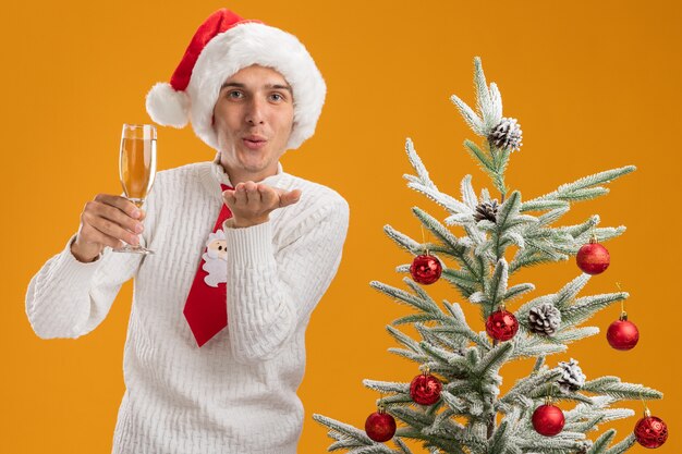 Pleased young handsome guy wearing christmas hat and santa claus tie standing near decorated christmas tree holding glass of champagne looking at camera sending blow kiss isolated on orange background