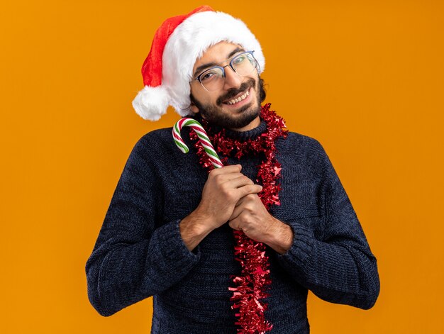 довольный молодой красивый парень в новогодней шапке с гирляндой на шее держит рождественские конфеты на оранжевой стене
