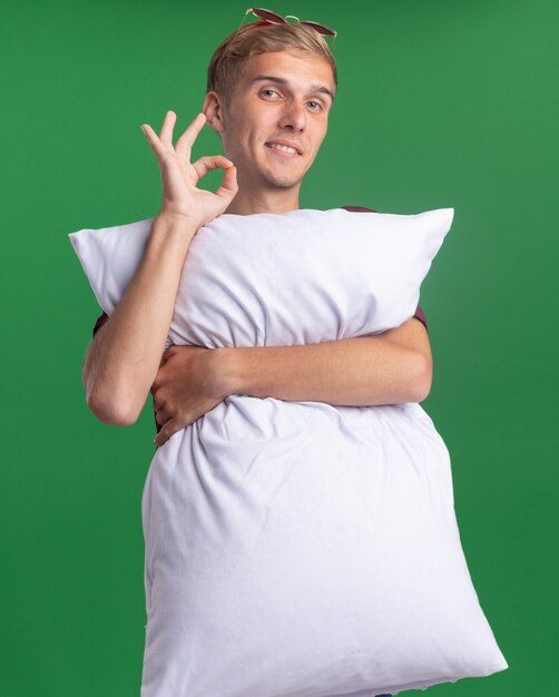 Pleased young handsome guy wearing red shirt hugged pillow showing okay gesture isolated on green wall
