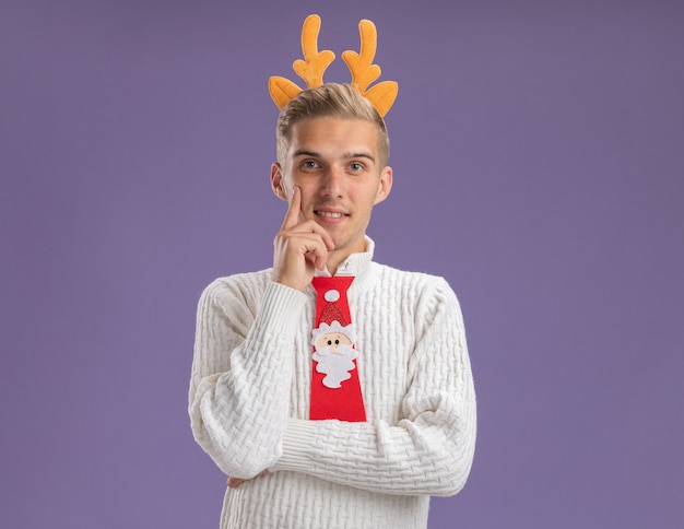 Free photo pleased young handsome guy wearing reindeer antlers headband and santa claus tie looking at camera touching chin isolated on purple background with copy space
