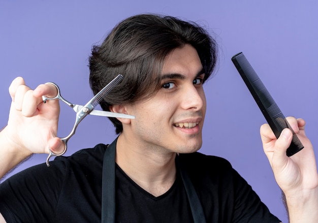Pleased young handsome male barber in uniform holding scissors and comb isolated on blue