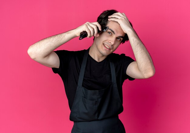 Pleased young handsome male barber in uniform timming his hair with hair clippers isolated on pink