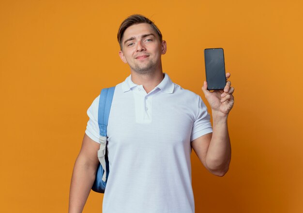 Pleased young handsome male student wearing back bag holding phone isolated on orange