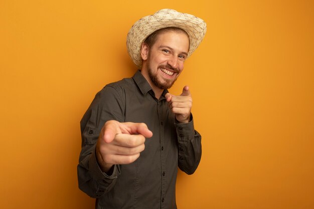 Pleased young handsome man in grey shirt and summer hat pointing with index fingers at camera smiling cheerfully 