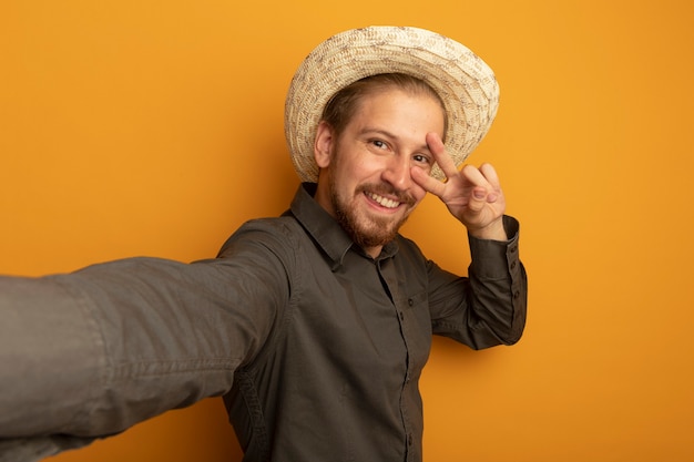 Pleased young handsome man in grey shirt and summer hat smiling cheerfully showing v-sign 