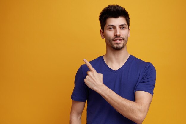 Pleased young handsome man looking at camera pointing to side on yellow background with copy space