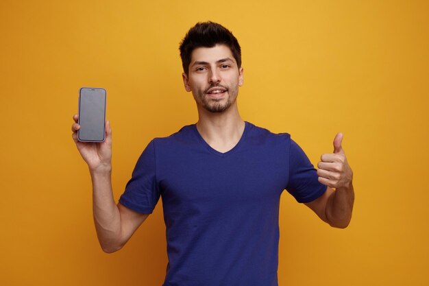 Довольный молодой красивый мужчина смотрит в камеру, показывает мобильный телефон на камеру и показывает большой палец вверх на желтом фоне