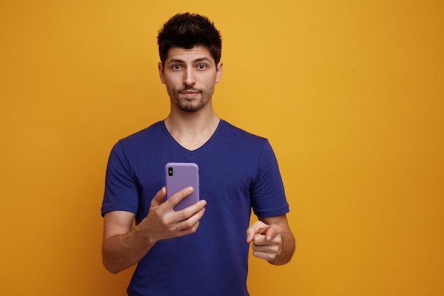 Pleased young handsome man pointing and looking at camera stretching mobile phone out towards camera on yellow background