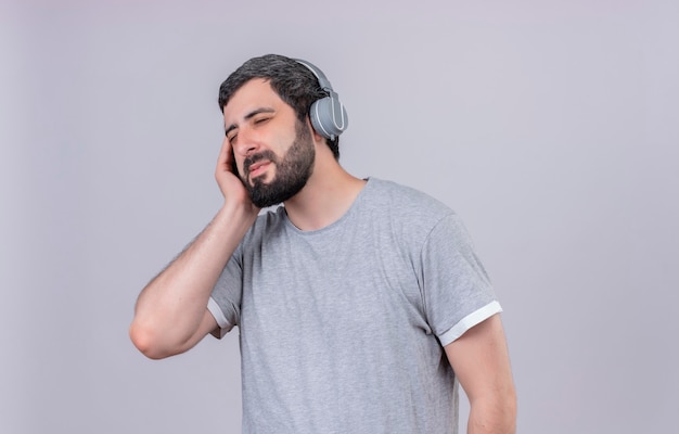 Pleased young handsome man wearing headphones listening to music with hand on headphone and closed eyes isolated on white wall