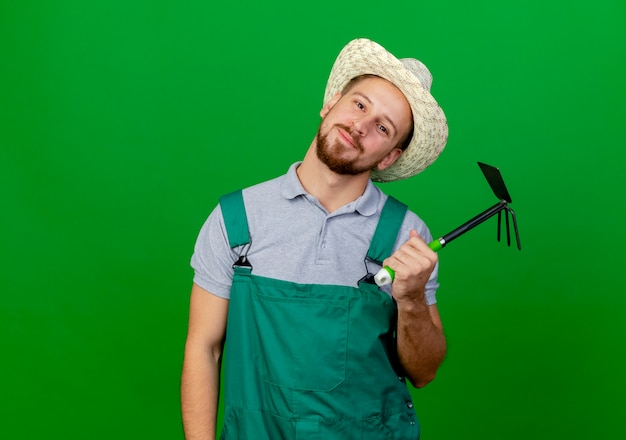 Pleased young handsome slavic gardener in uniform and hat holding hoe-rake looking 