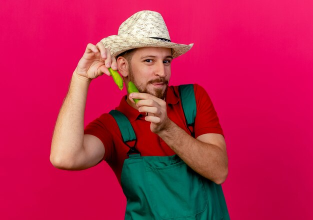 Free photo pleased young handsome slavic gardener in uniform and hat holding pepper halves looking