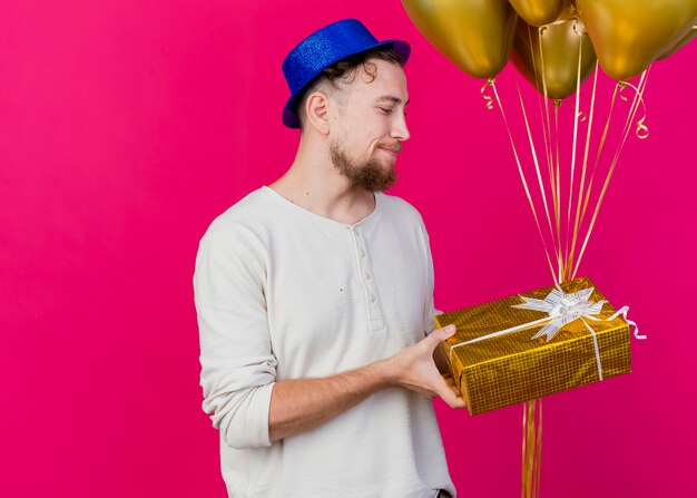 Pleased young handsome slavic party guy wearing party hat holding balloons and gift box looking at gift box isolated on crimson background with copy space
