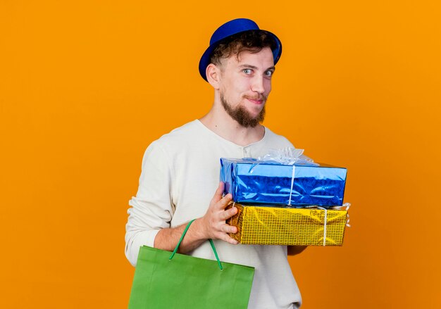 Pleased young handsome slavic party guy wearing party hat holding gift boxes and paper bag looking at camera isolated on orange background with copy space