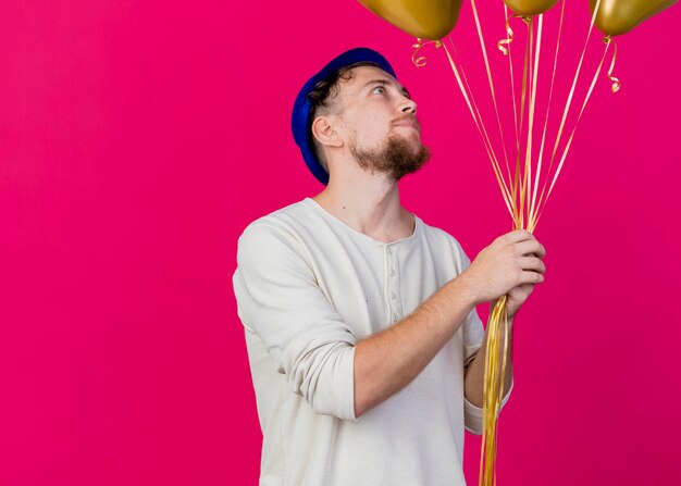 Soddisfatto giovane ragazzo slavo bello del partito che indossa il cappello del partito che tiene e che guarda gli aerostati isolati sulla parete rosa con lo spazio della copia